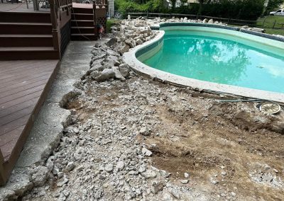 Broken concrete and dirt around a backyard pool under renovation, next to a wooden deck.