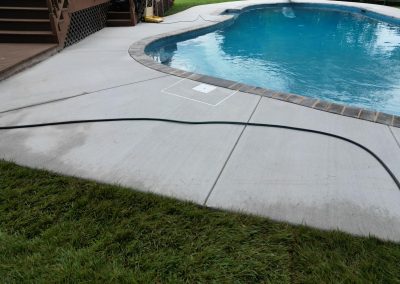 A garden hose lies on concrete next to a swimming pool and grassy lawn.