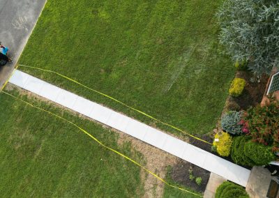 Aerial view of a freshly poured sidewalk bordered by yellow tape on a green lawn.