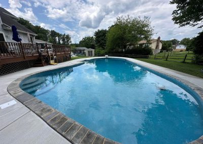 A backyard swimming pool with clear blue water, surrounded by trees, grass, and a wooden deck.