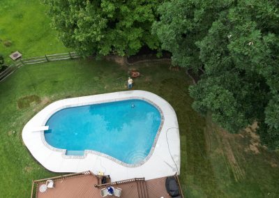 Aerial view of a backyard pool surrounded by grass, trees, and a wooden deck.