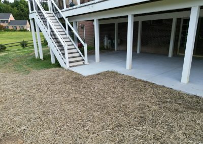 A house with a deck, stairs, concrete patio, and straw spread over the yard near the patio.