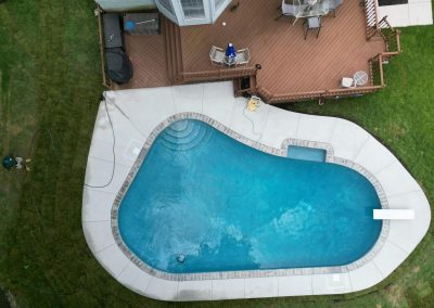 Aerial view of a backyard pool, deck with furniture, and two people working near the pool and on the grass.