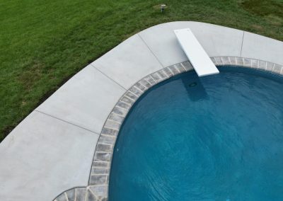 A backyard pool with a diving board, surrounded by a concrete deck and green grass.