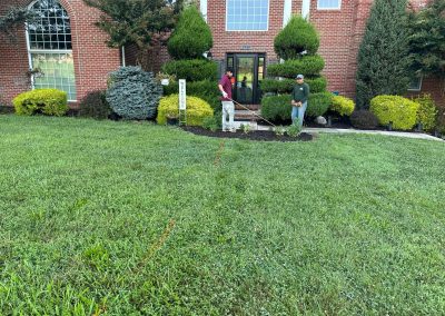 Two people stand in front of a building, marking a line on the grass in a landscaped yard.