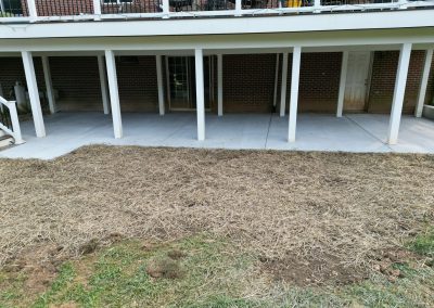 Covered concrete patio with white columns; straw-covered ground in foreground and brick wall in background.