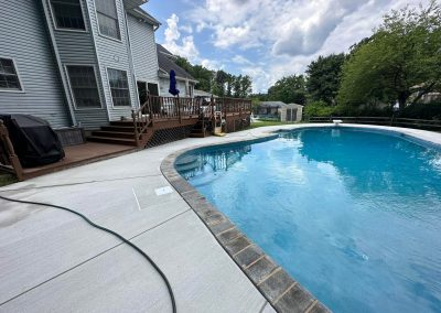 Backyard pool with a wooden deck, grill, and patio furniture beside a gray house on a partly cloudy day.