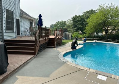 Two people clean a backyard pool next to a wooden deck on a sunny day.