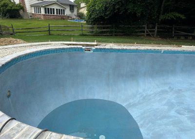 Empty in-ground swimming pool under construction in a backyard with a house and wooden fence in the background.