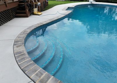 Curved pool steps leading into a clean, blue swimming pool next to a concrete patio and wooden deck.