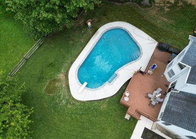 Aerial view of a backyard with a swimming pool, wooden deck, patio furniture, and surrounding green lawn.