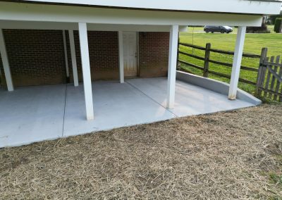 A concrete patio under a raised deck, beside a wooden fence and a grassy yard.