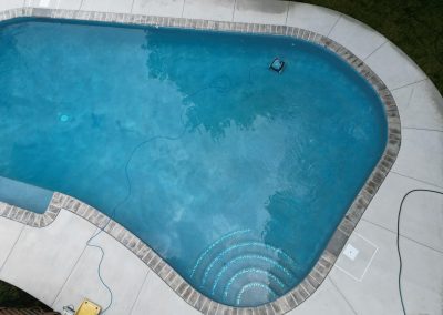 A clean, blue swimming pool with a pool cleaner and steps, surrounded by concrete decking.