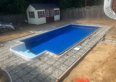 Rectangular in-ground pool with blue water; concrete forms and gravel surround it, shed in background.