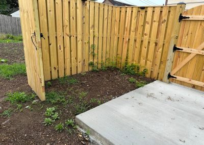 Wooden fence corner with small weeds and plants growing on soil next to a concrete patio.