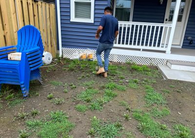 A man walks on a patchy, grassy yard in front of a blue house with a porch and stacked blue chairs nearby.