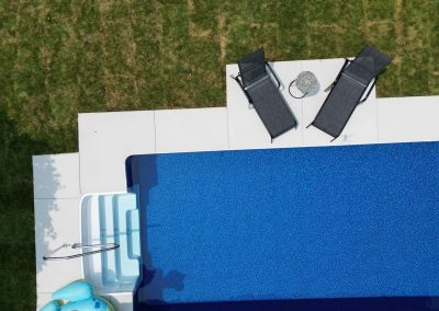 Aerial view of two lounge chairs by a blue swimming pool and a small round table on a grassy lawn.
