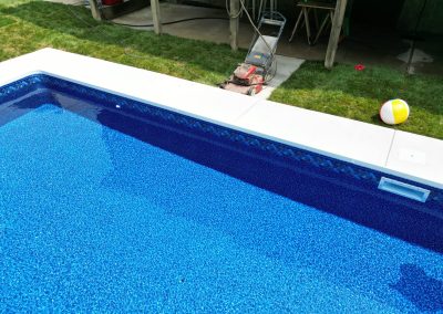 A lawn mower and a beach ball sit on grass beside a backyard swimming pool.