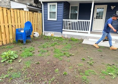 A man walks past patchy grass and weeds in a yard beside a house with blue siding and a white porch.