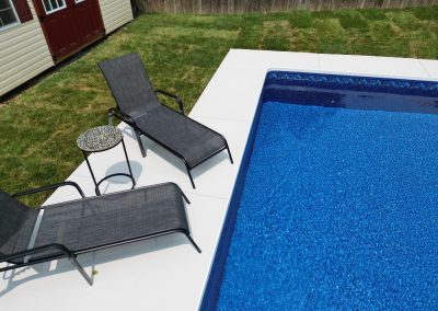 Two black lounge chairs and a small table sit beside a blue swimming pool on a concrete patio.