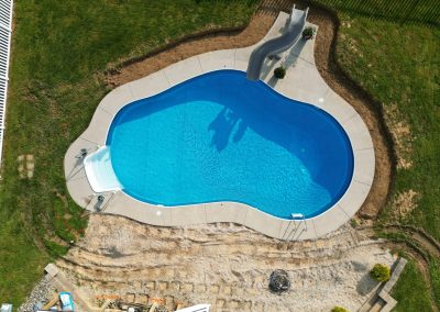 Aerial view of a backyard swimming pool with a slide, surrounded by grass and a dirt area.