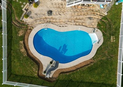 Aerial view of a backyard swimming pool with a slide, surrounded by grass and a white fence.