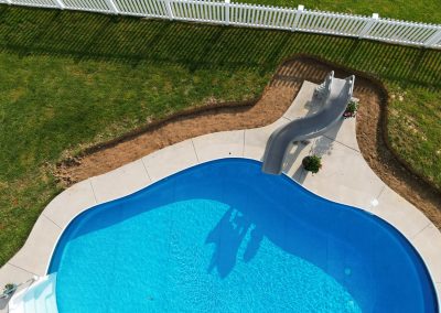 A backyard pool with a curved slide, surrounded by grass and a white fence on a sunny day.