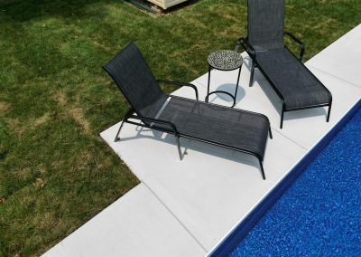 Two black lounge chairs and a small round table sit beside a bright blue pool on a concrete patio.