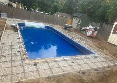 A rectangular in-ground pool under construction, surrounded by gravel and metal rebar in a fenced backyard.