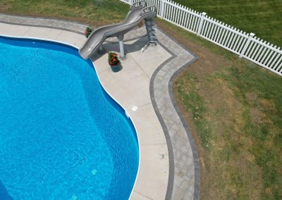 A backyard pool with a gray slide, curved patio, white fence, and potted plants beside the water.