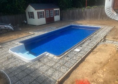 A rectangular in-ground pool under construction, surrounded by wire mesh and gravel, with a shed in the background.
