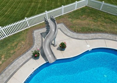 A curved gray pool slide beside a blue swimming pool, surrounded by a white fence and green grass.