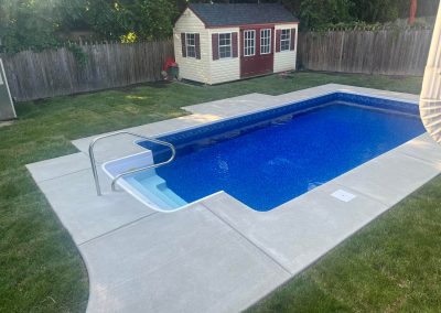 Backyard with an in-ground swimming pool, concrete deck, grass, and a small shed near a wooden fence.