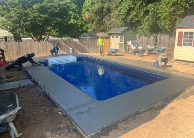 Workers smooth fresh concrete around a backyard swimming pool on a sunny day, with sheds and trees nearby.