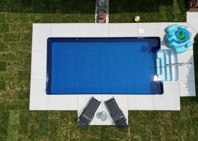 A rectangular backyard pool with two lounge chairs, a small table, and a blue pool float beside the steps.