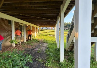 Two people in orange shirts work near the brick wall under a wooden deck, with plants and stairs visible.