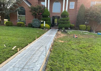 A new concrete walkway is being constructed leading to the entrance of a red brick house.