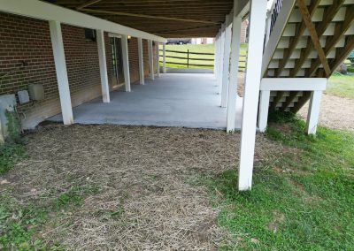 Under-deck patio with concrete floor, wooden posts, stairs, and adjacent patch of straw and grass.