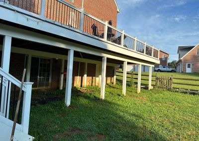 View of a brick house with a raised deck, white railings, and grassy backyard on a sunny day.