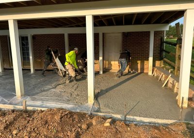 Four workers spread and smooth wet concrete under a covered patio next to a brick building.