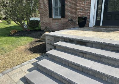 Stone steps lead up to a front porch of a brick house, next to landscaped grass and mulch.