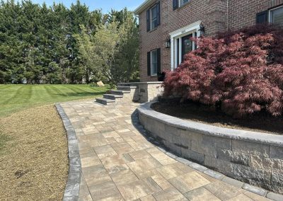 Paved stone walkway curving toward brick house, steps, red-leaf shrub, and neatly mowed lawn.