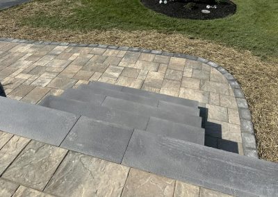 Gray stone steps lead down to a paved patio bordered by grass and a mulched area with rocks.