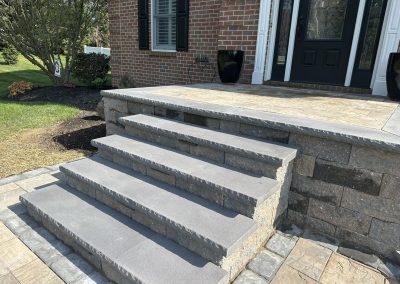 Stone steps leading to a front porch with a black door and potted plants, next to a green lawn.