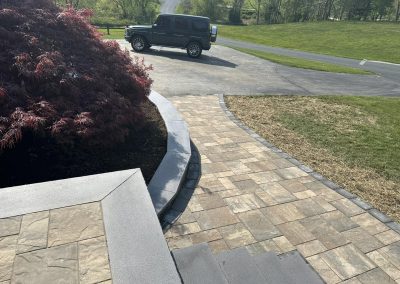 Stone walkway leading to a driveway with a parked black SUV, next to grass and a red-leafed shrub.