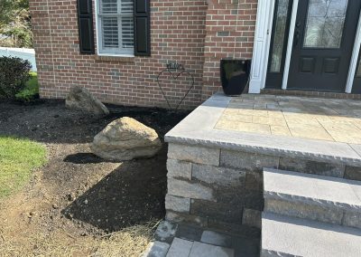Large rocks and bare soil by a brick house entrance with stone steps and a heart-shaped garden sign.