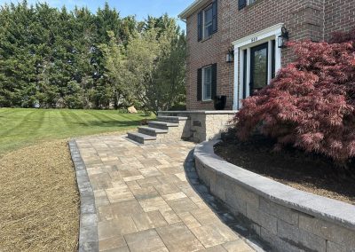 Brick house with a stone walkway, steps, red tree, and manicured lawn bordered by tall greenery.