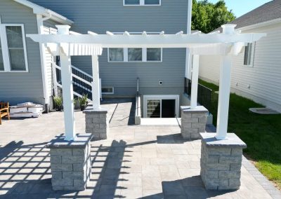 White pergola with stone pillars on a spacious patio beside a gray house and green lawn.