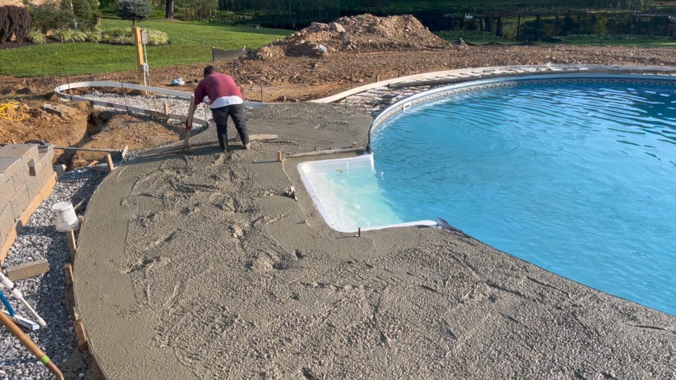 Person smoothing wet concrete around a backyard pool under construction on a sunny day.