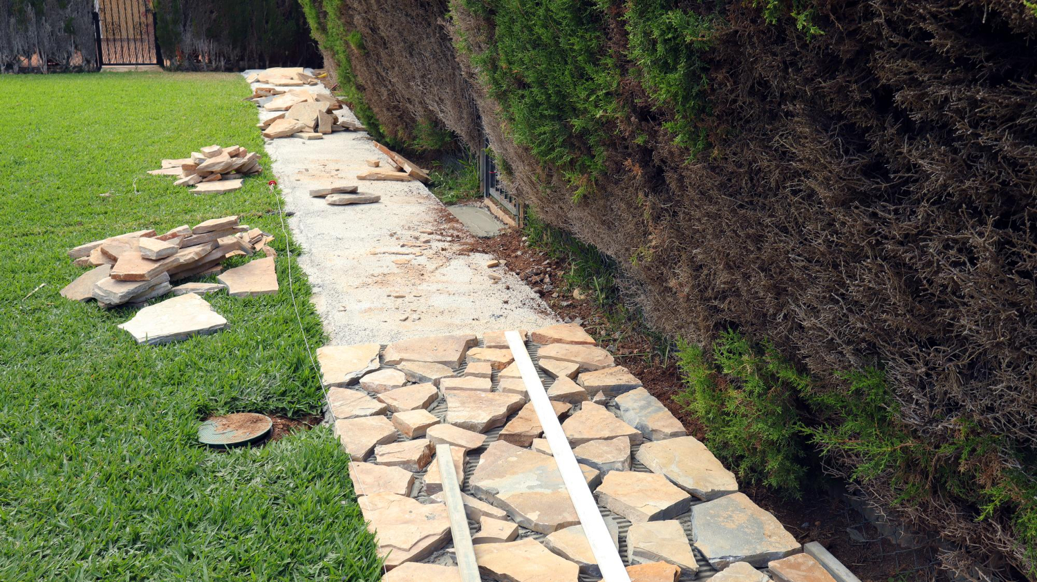 10290 Stone pathway under construction beside a hedge, with paving stones and tools scattered on the grass.